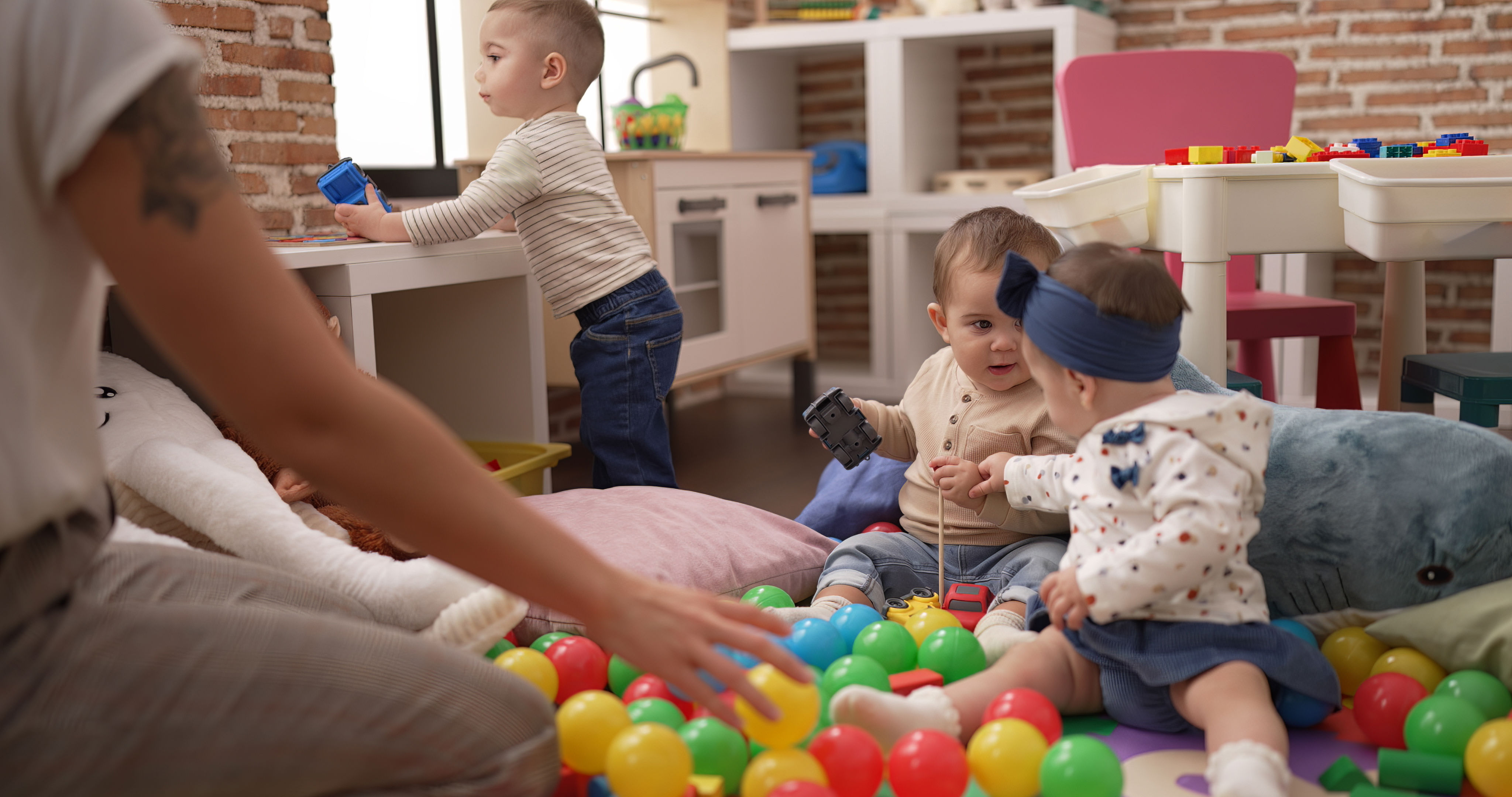 To blebørn leger på gulvet mellem små bolde - et lidt større barn står op og der sidder en voksen overfor dem