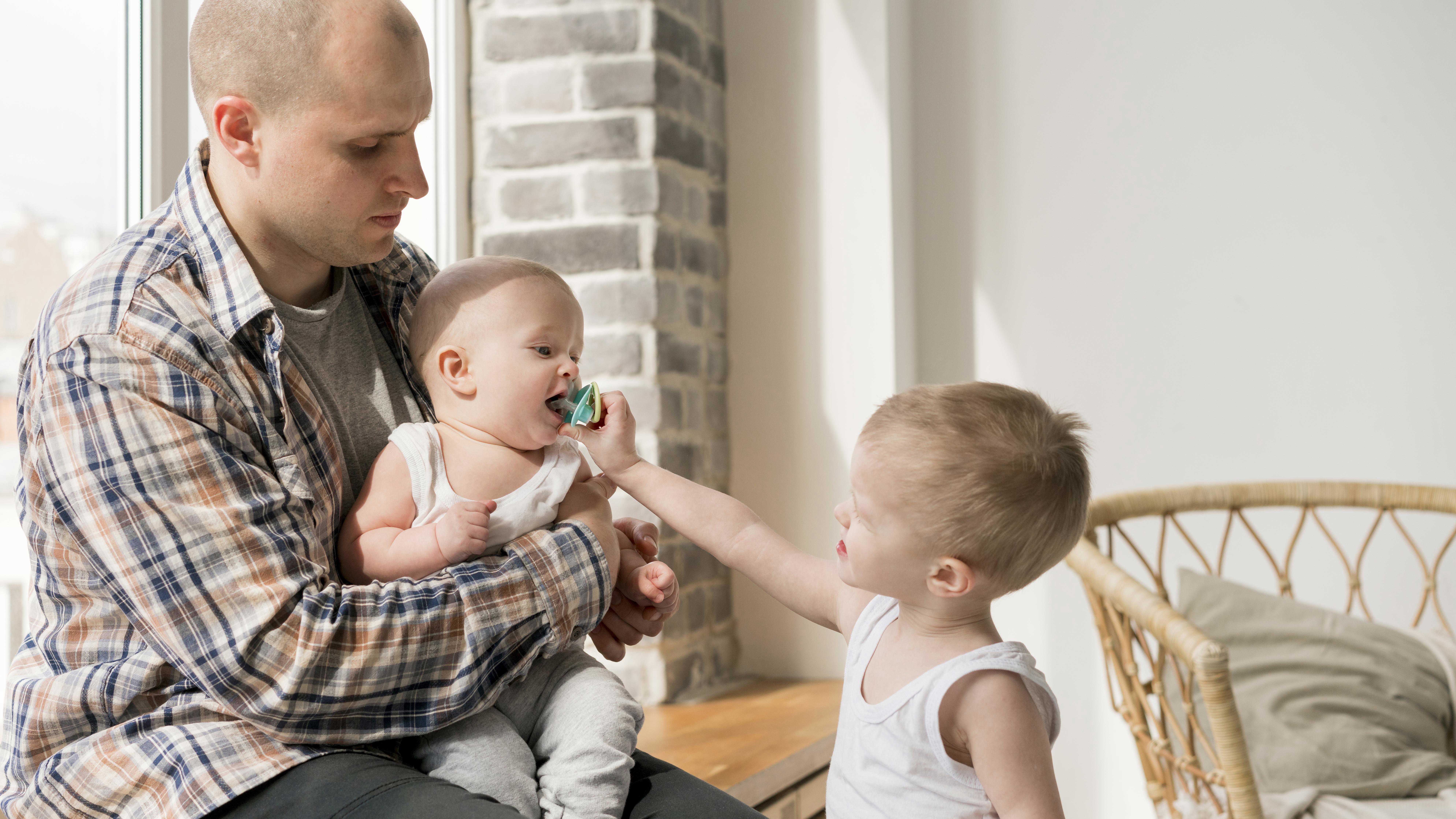 Stående dreng giver baby, der sidder hos far, sut i munden