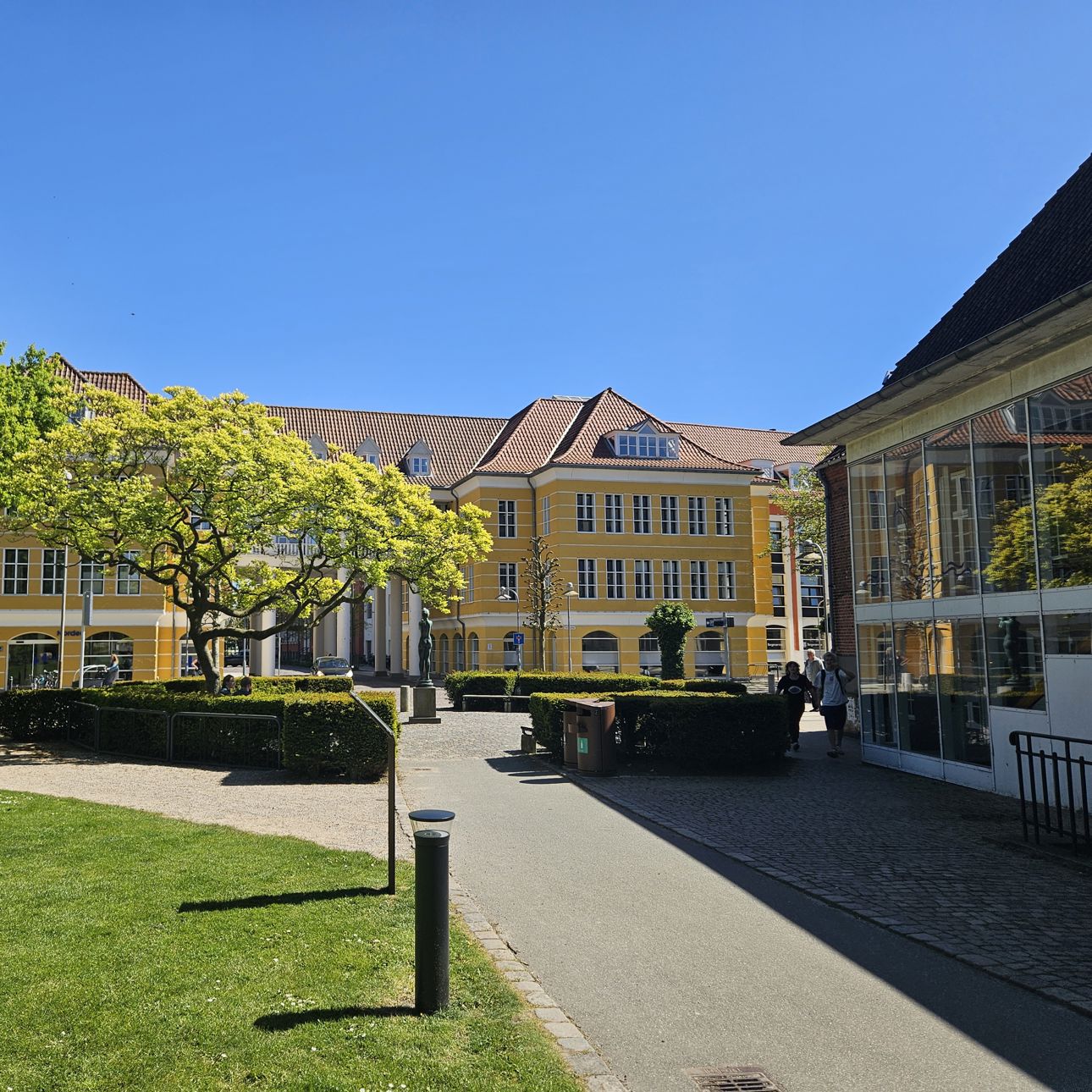 En sti med græs omkring, fører ned til en stor gul bygning med hvide vinduer. Foran bygningen er en plads omkranset af en rund hæk og et stort træ.