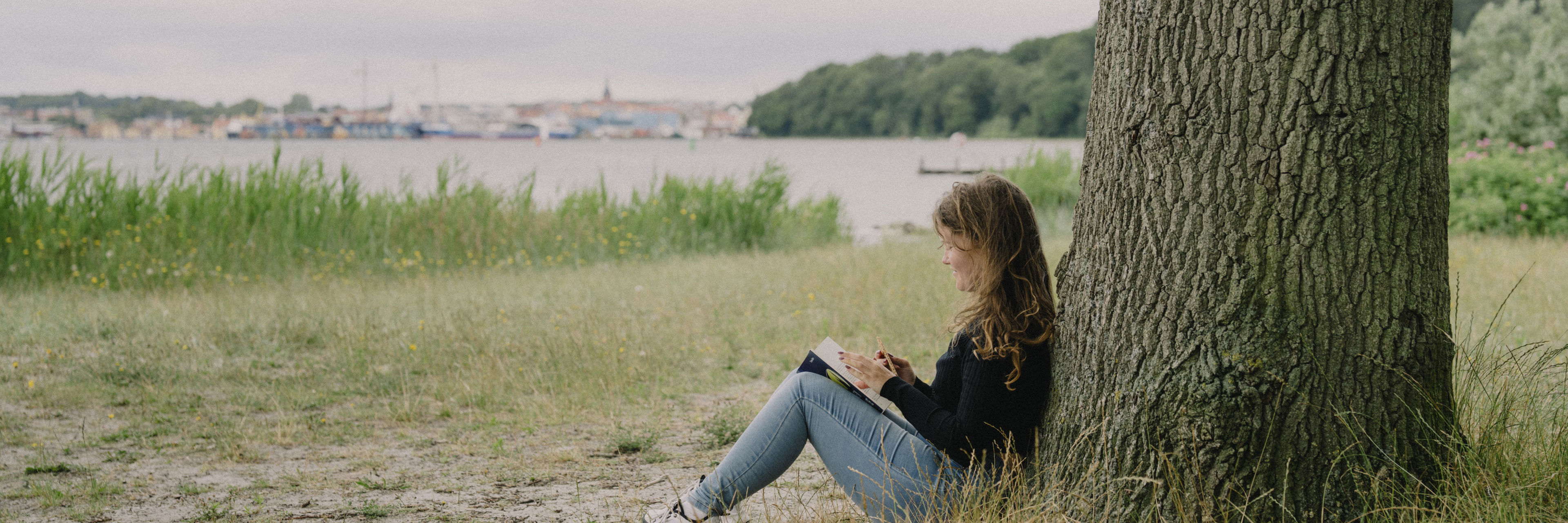 Pige sidder op ad træ og læser bog - i baggrunden Svendborg By set fra vandsiden