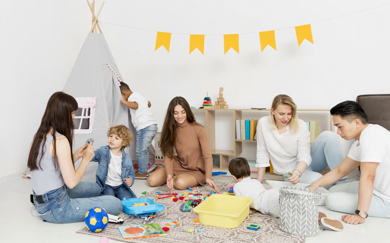 3 kvinder og en mand sidder på gulvet og leger med tre børn, hvoraf det ene barn kigger ind i et lille tipitelt
