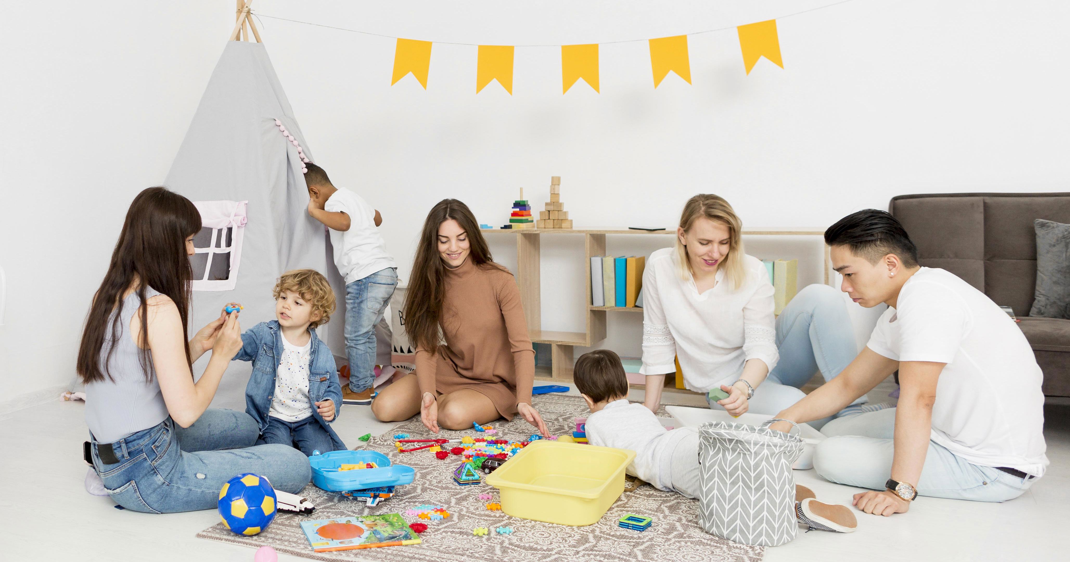 3 kvinder og en mand sidder på gulvet og leger med tre børn, hvoraf det ene barn kigger ind i et lille tipitelt