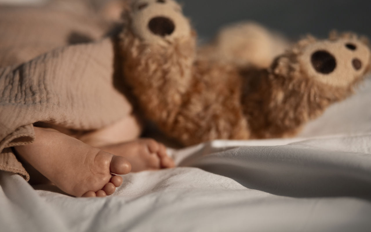Baby fødder der stikker ud under tæppe. Brun bamses fødder ligger og stritter i vejret ved siden af baby.