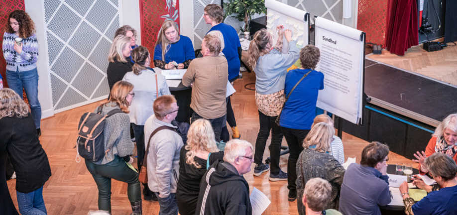 Mange mennesker samlet i et stort lokale. Nogle står og snakker ved caféborde, mens andre hænger post its på et lærred.