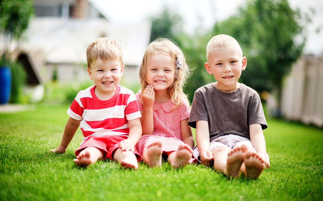 Tre mindre børn sidder på græsset ved siden af hinanden