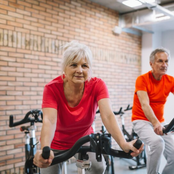 En ældre kvinde sidder på en motionscykel. En ældre mand på en motionscykel ses i baggrunden