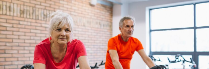 En ældre kvinde sidder på en motionscykel. En ældre mand på en motionscykel ses i baggrunden