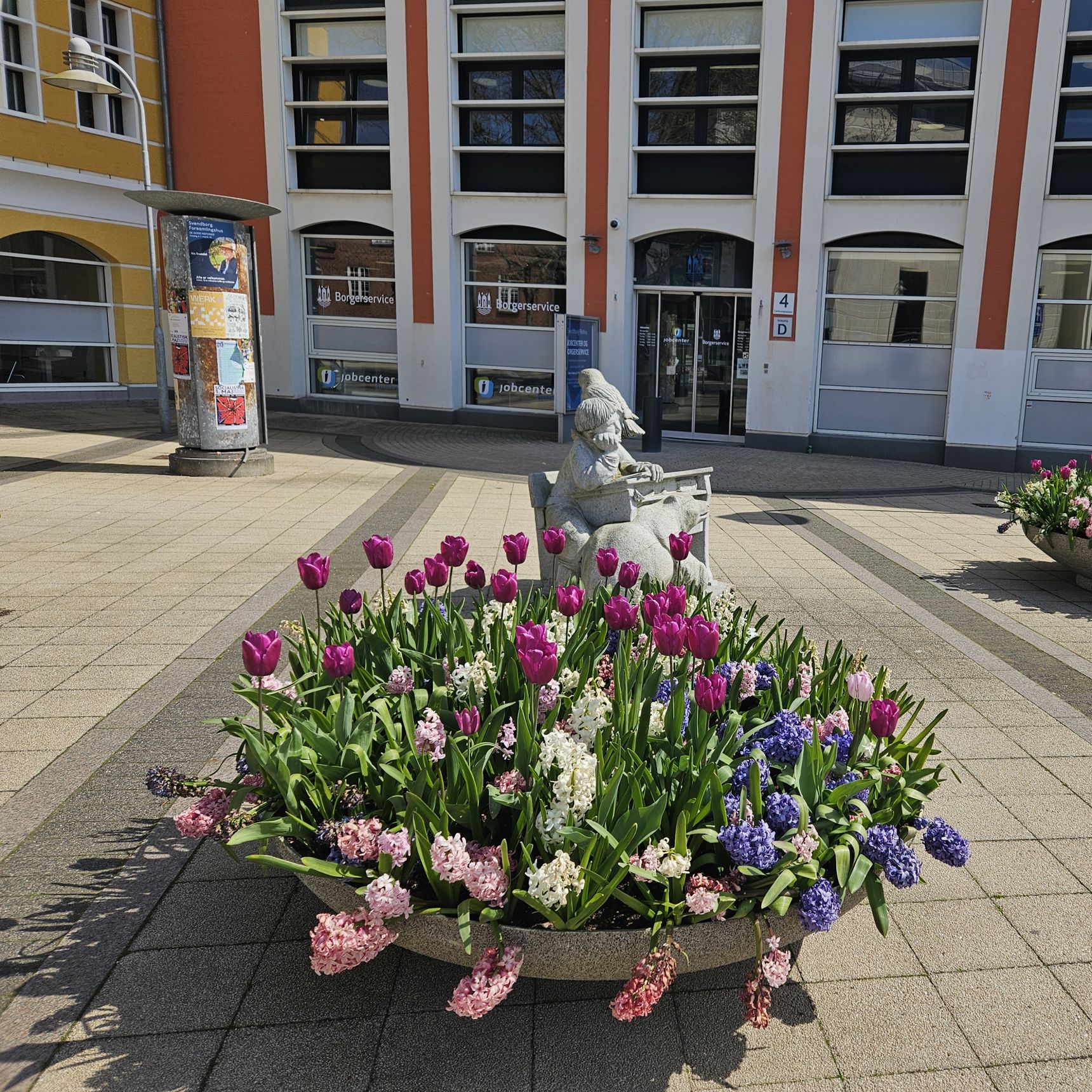 En kumme med forårsblomster foran indgangen til en stor bygning, der er hvid og rød.