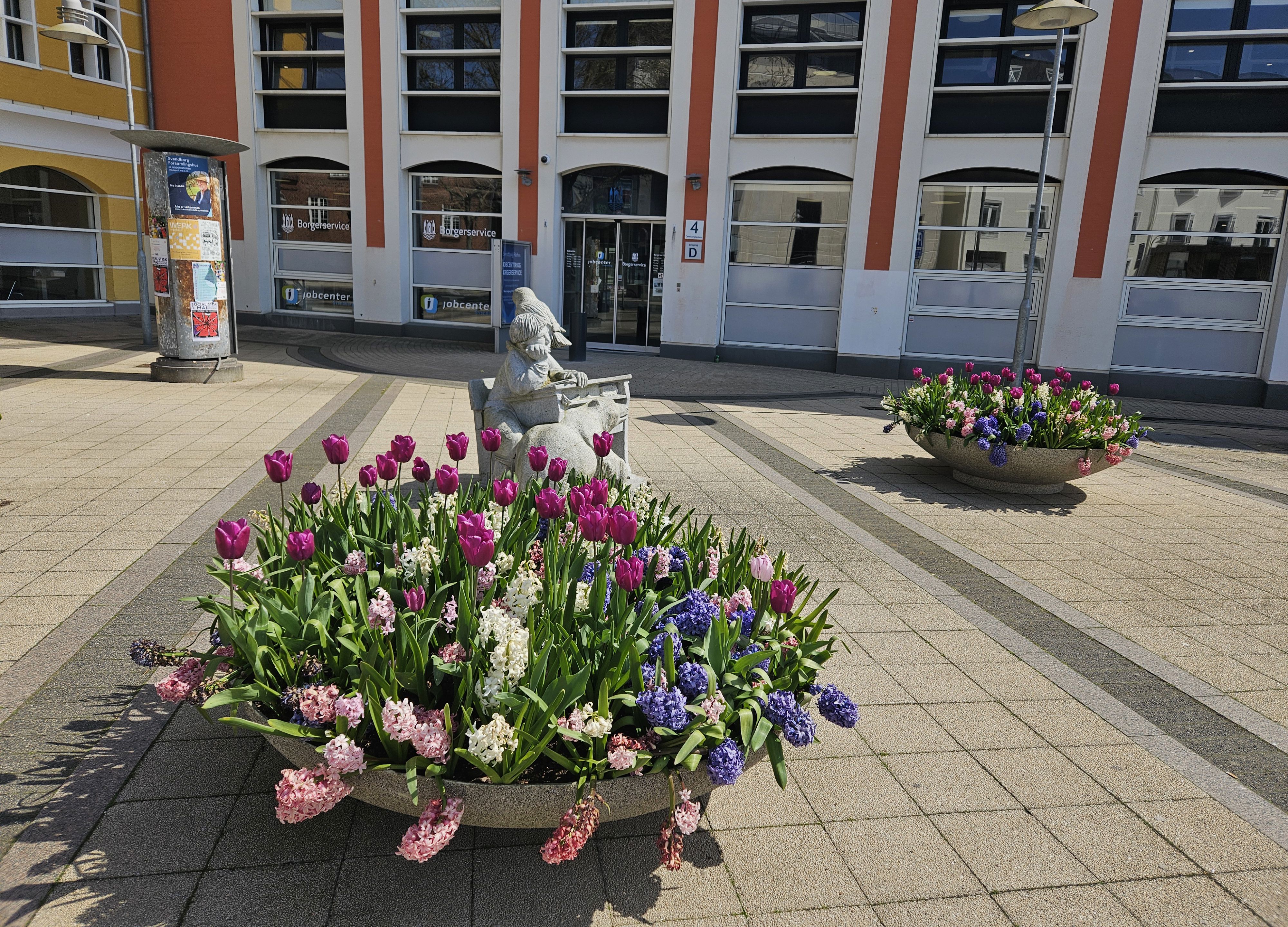 En kumme med forårsblomster foran indgangen til en stor bygning, der er hvid og rød.