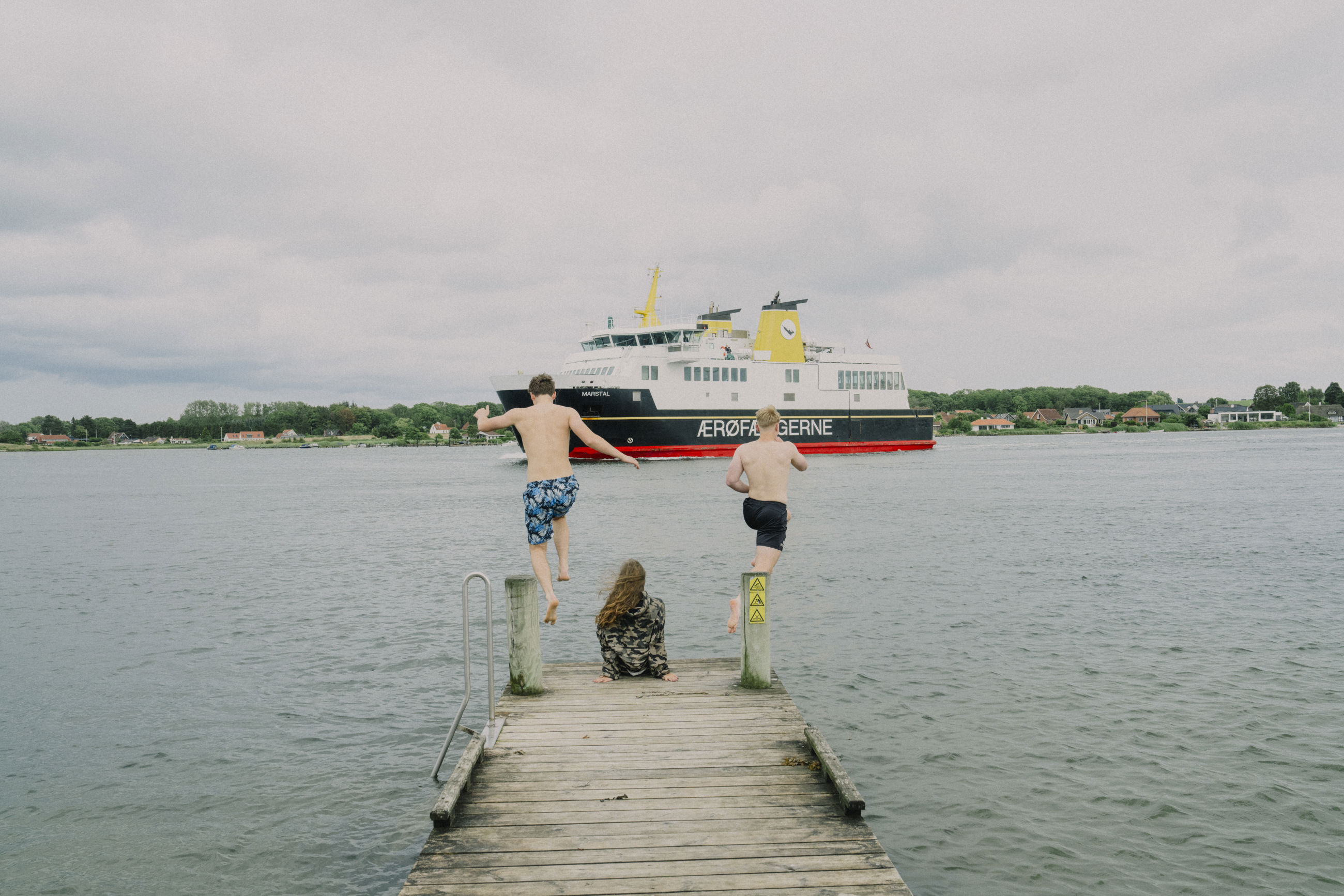 To unge springer i vandet fra badebro imens ærø-færgen sejler forbi