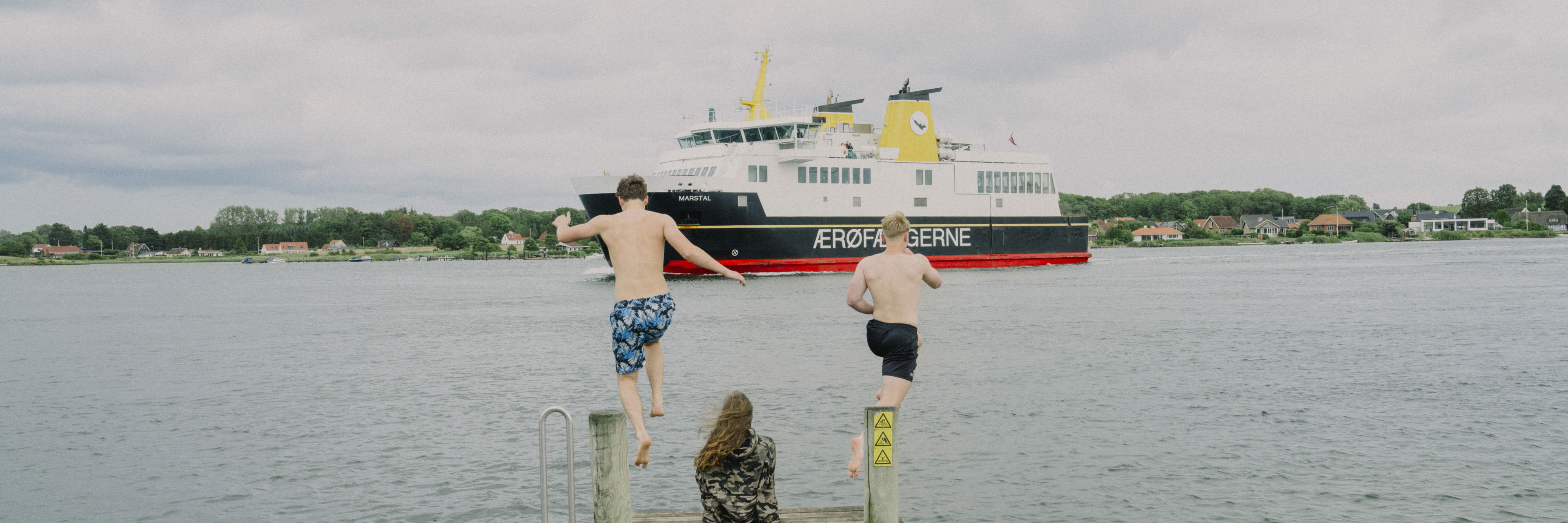 To unge springer i vandet fra badebro imens ærø-færgen sejler forbi