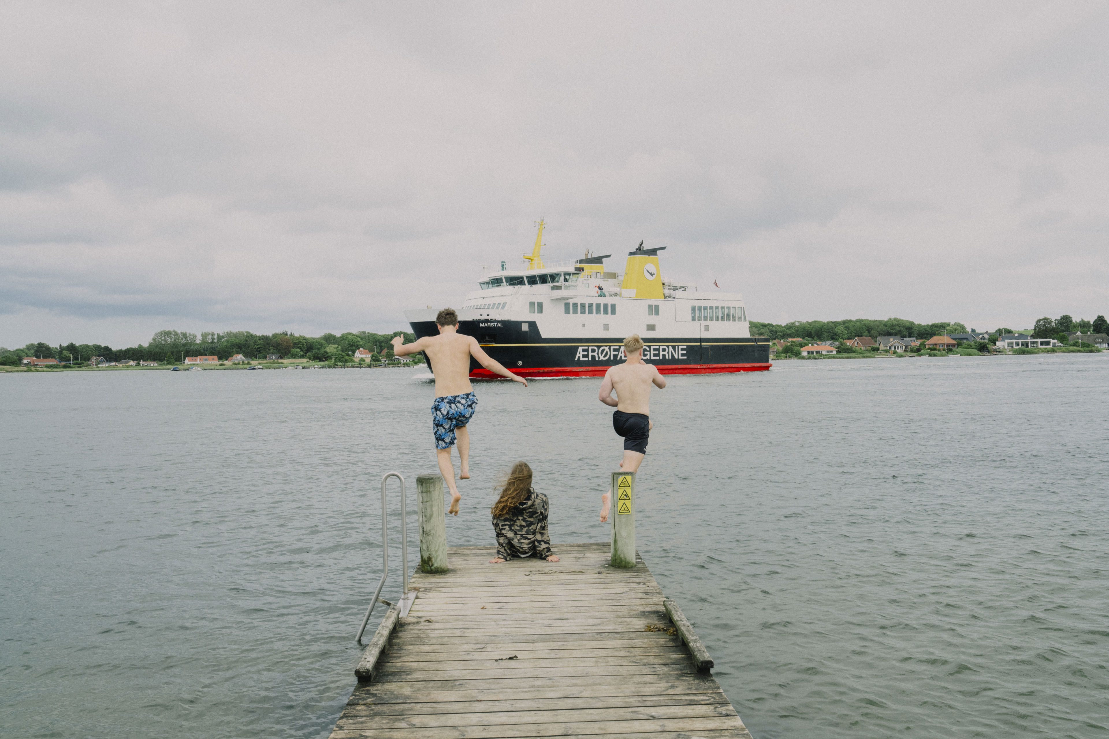 To unge springer i vandet fra badebro imens ærø-færgen sejler forbi