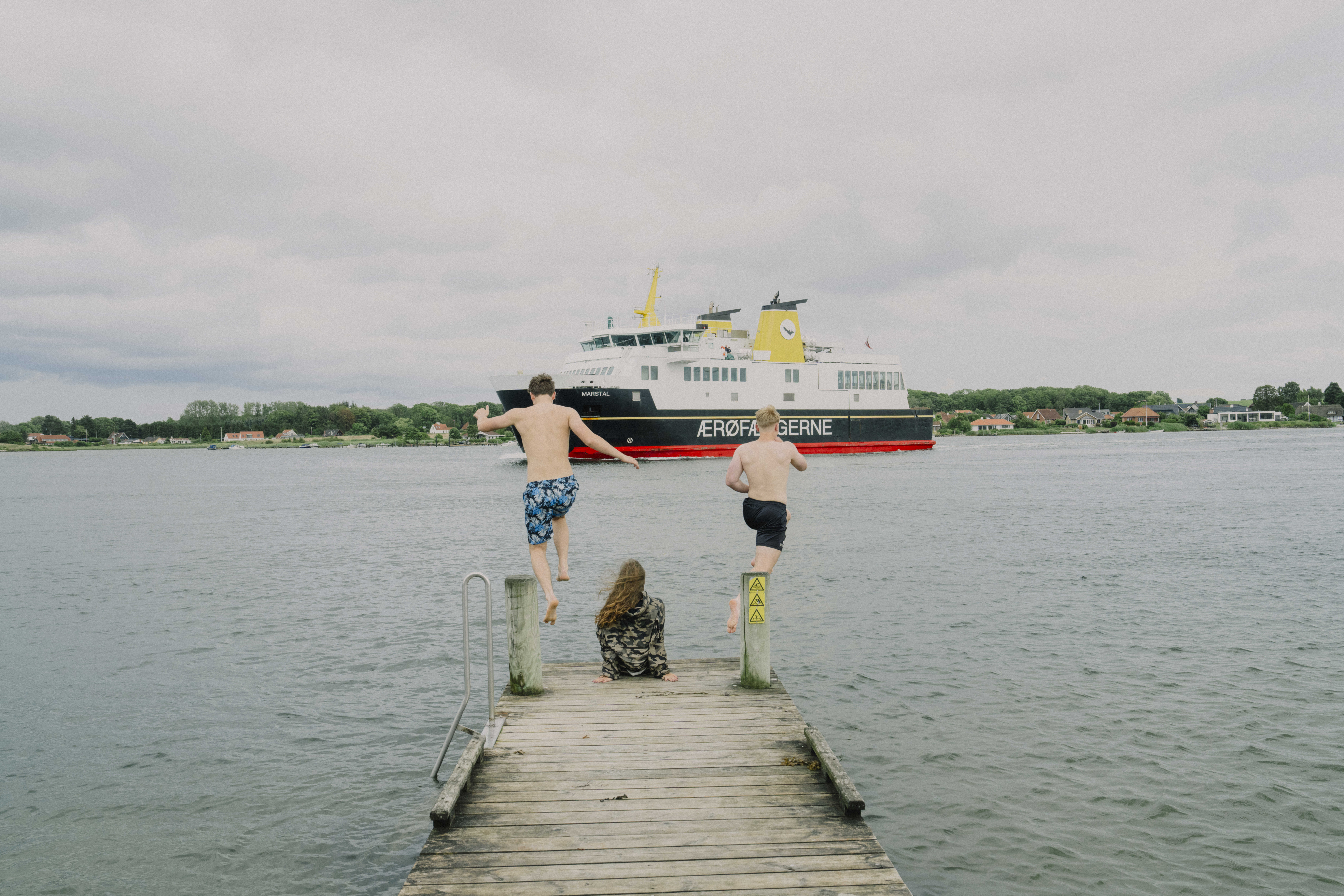 To unge springer i vandet fra badebro imens ærø-færgen sejler forbi