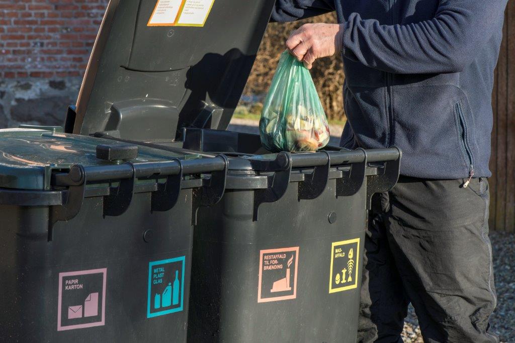 To sorte skraldespande med piktogrammer for affaldssortering og en person, der fylder en pose i en af spandene