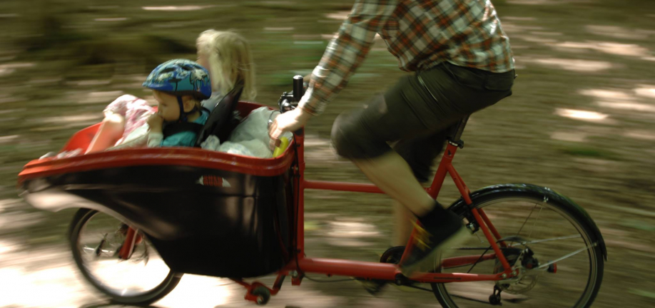 Close up foto af ladcykel med to børn i ladet og en far, der cykler hurtigt forbi